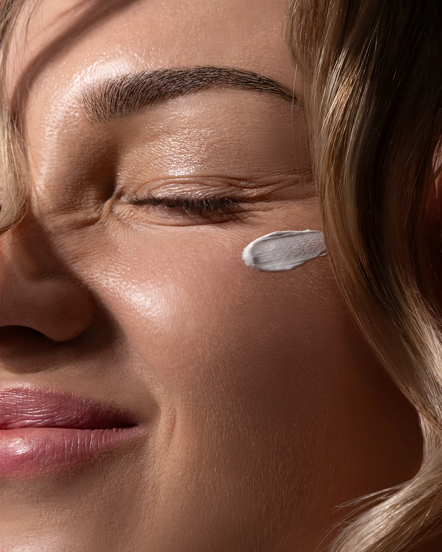 Close-up skincare commercial portrait of a smiling woman with a cosmetic cream swatch on her cheek, featuring Dodge & Burn, preserved natural skin texture and fine lines, precise highlight control, and warm color grading. Photography by Inga Cebotari.