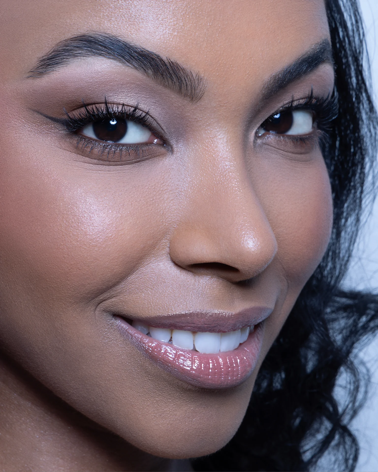 Close-up portrait of a smiling woman with dark skin, expertly retouched to showcase clean, textured skin and perfect makeup detail. Features include defined brows, precise eyeliner, polished teeth, and lip gloss, achieved through advanced dodge and burn, skin tone correction, and detail preservation. Photography by Inga Cebotari.